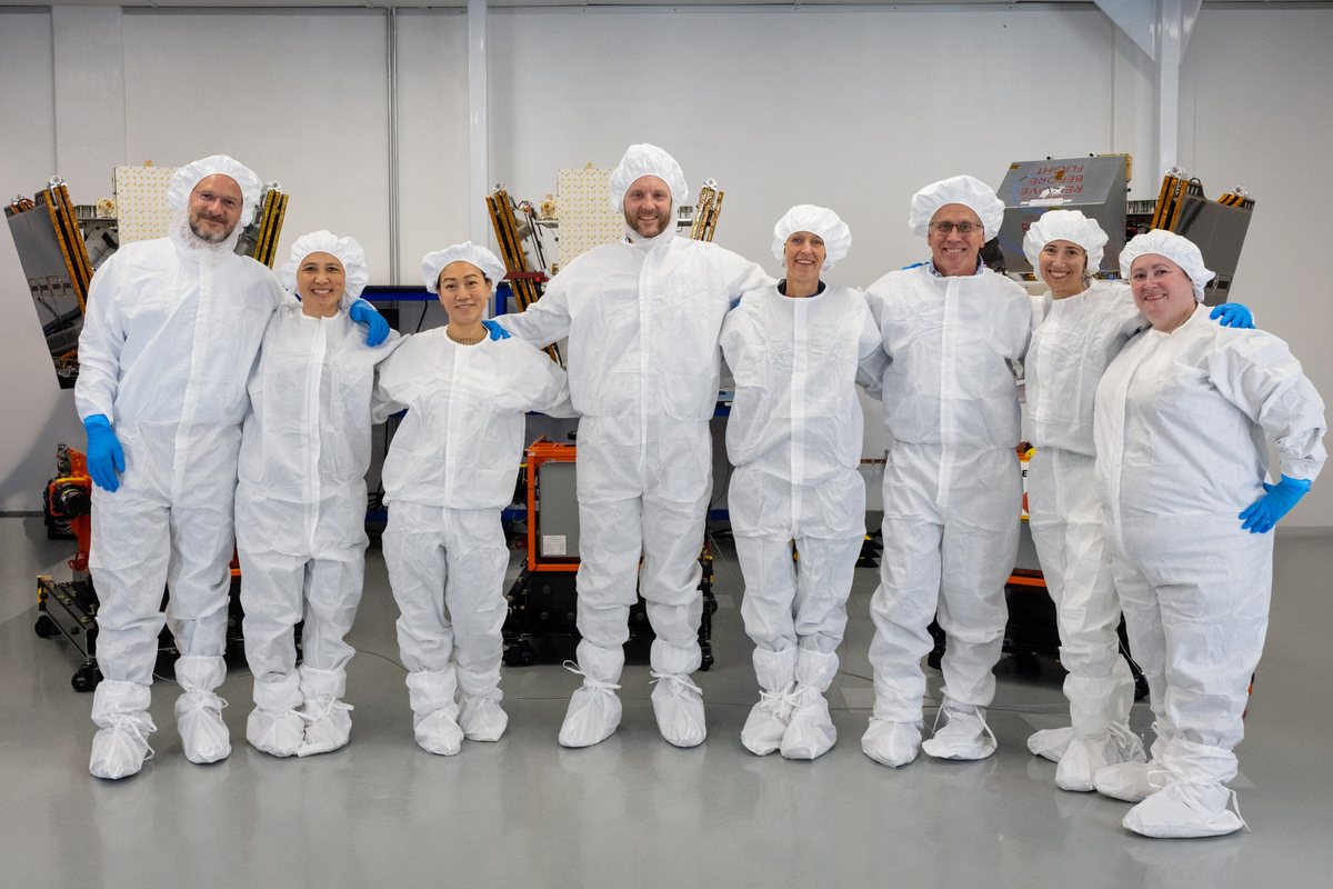 Eight people in white cleanroom suits, hairnets, and shoe covers stand arm in arm, smiling, in front of three FireSat satellites with gold-foil solar panels in a bright white assembly facility.